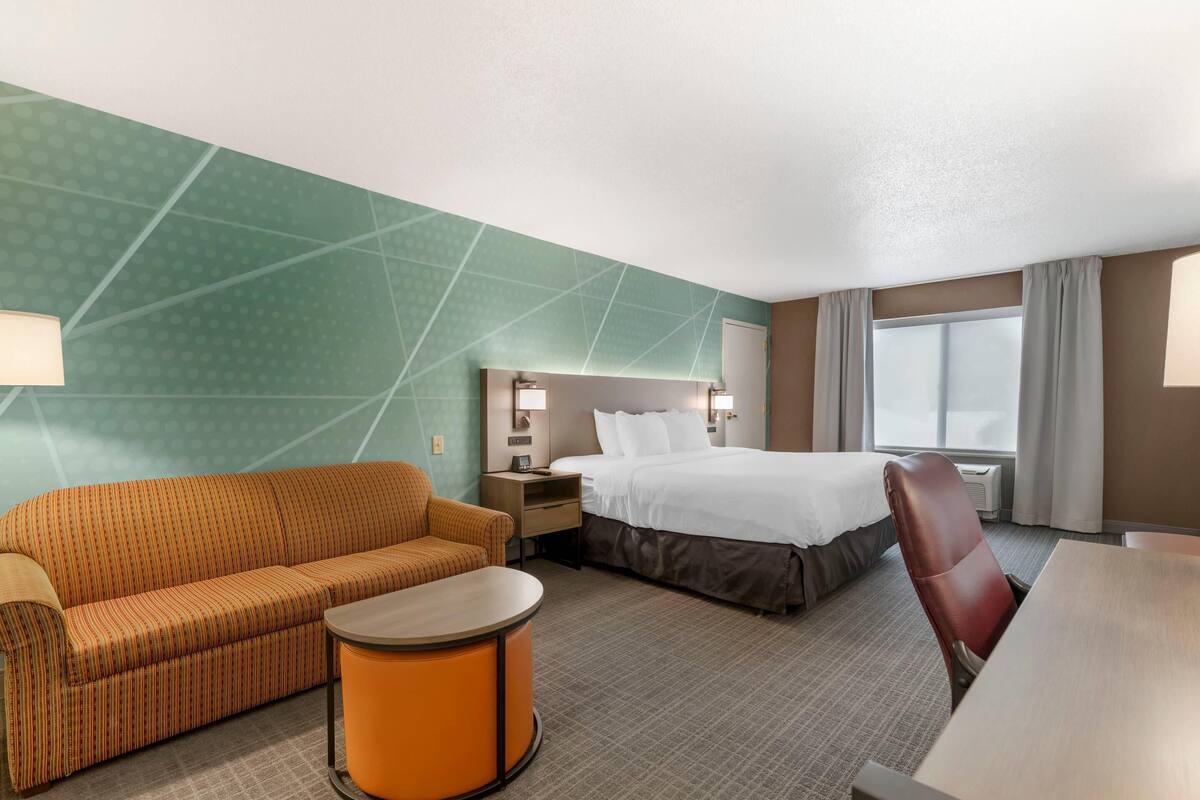 A hotel room with a bed, a sofa, a small table, a chair, a nightstand, and a lamp.