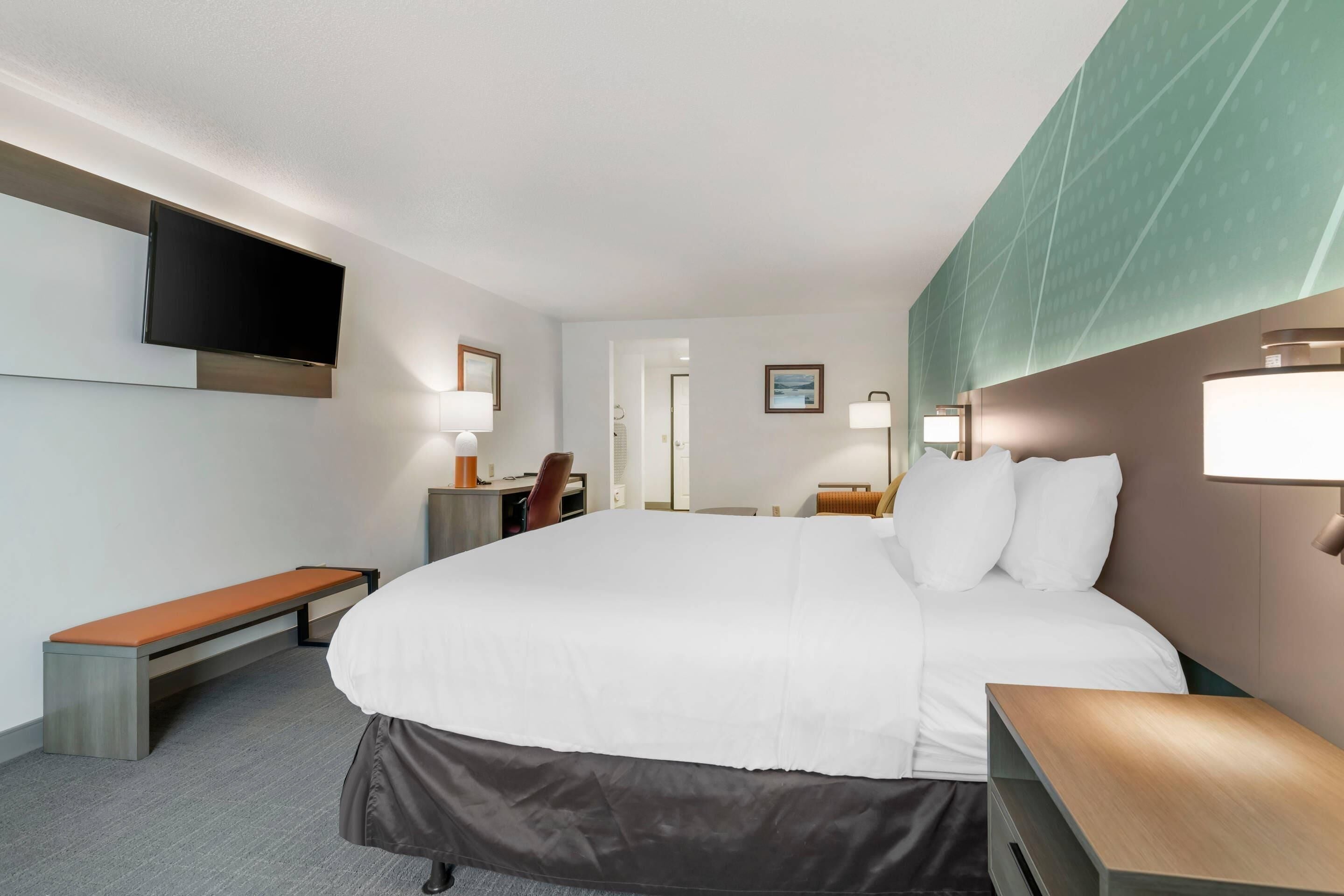 Ein Hotelzimmer mit einem großen Bett, Nachttischen, einer Bank, einem Flachbildfernseher und einem Schreibtisch mit Stuhl.
