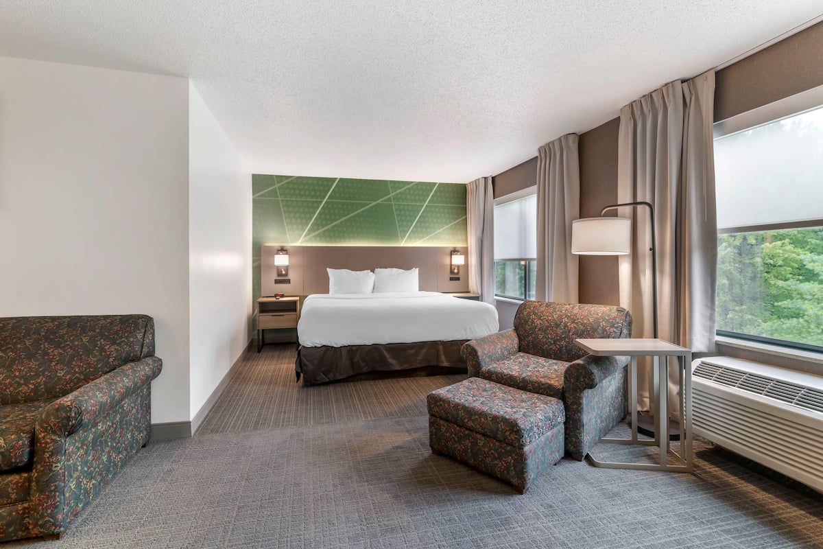 A hotel room with a bed, two armchairs, a small table, a nightstand, a lamp, and a window with a view.