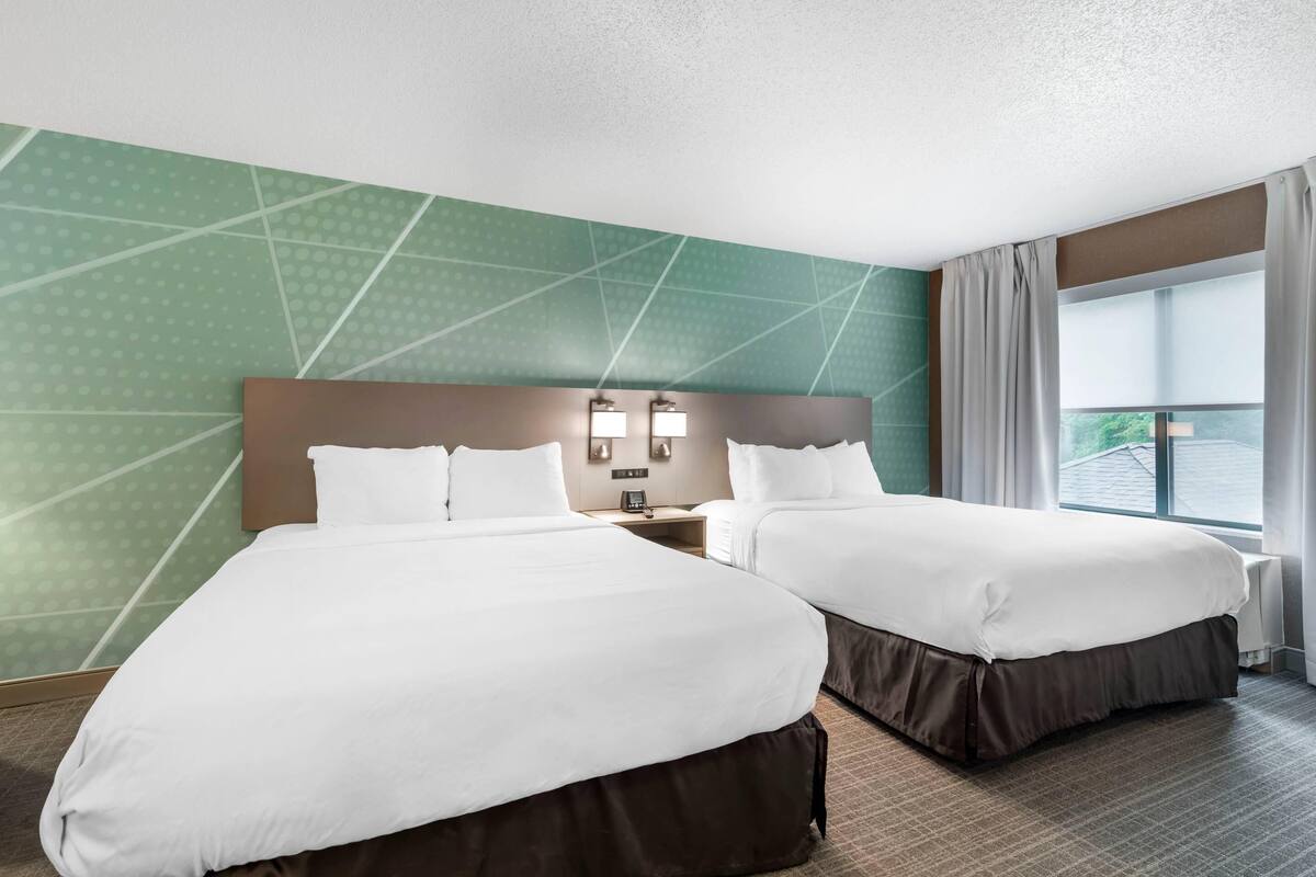 A hotel room with two beds, a nightstand, a wall-mounted lamp, and a window with curtains.