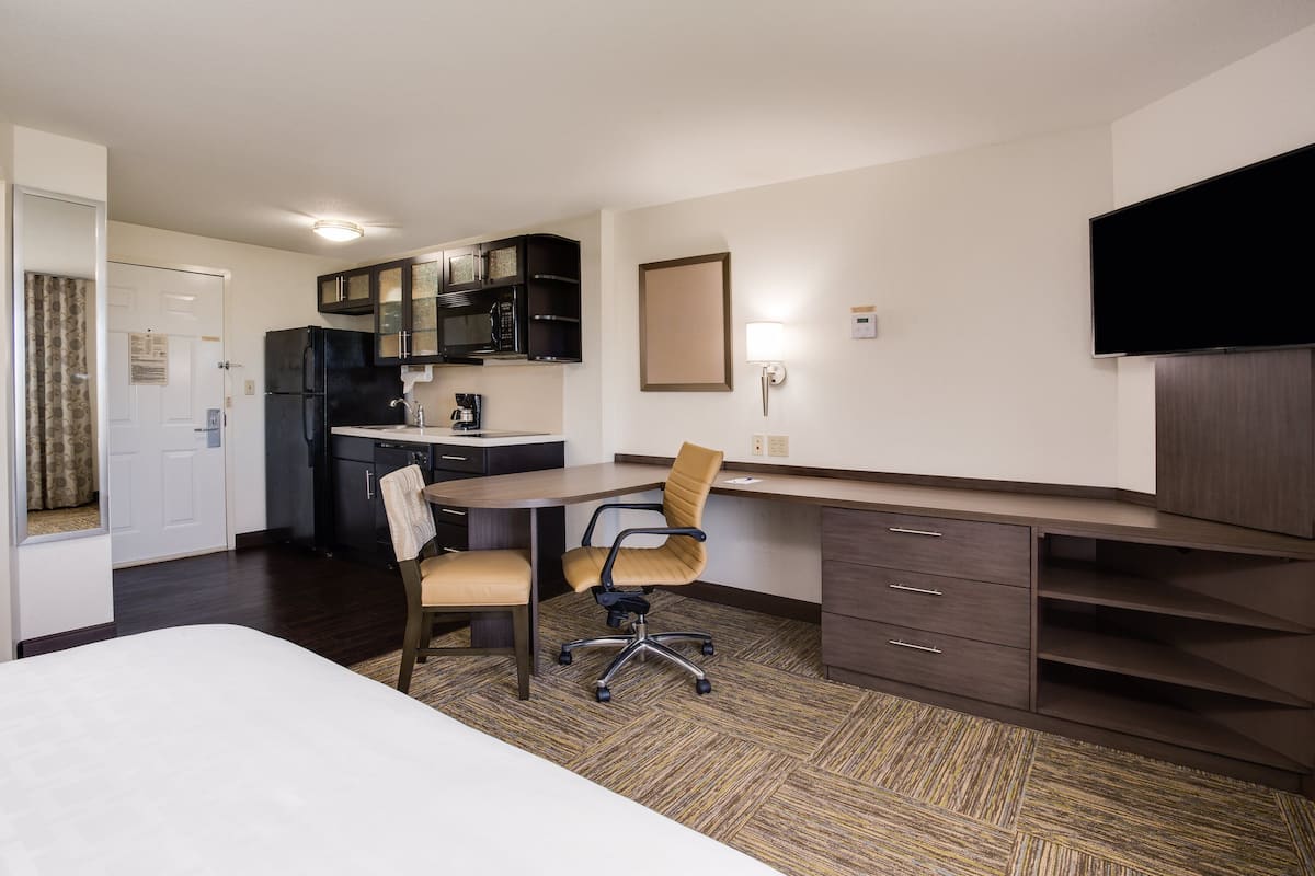 Une chambre d’hôtel compacte comprenant un lit, un bureau avec deux chaises, une kitchenette, un téléviseur à écran plat et un miroir.