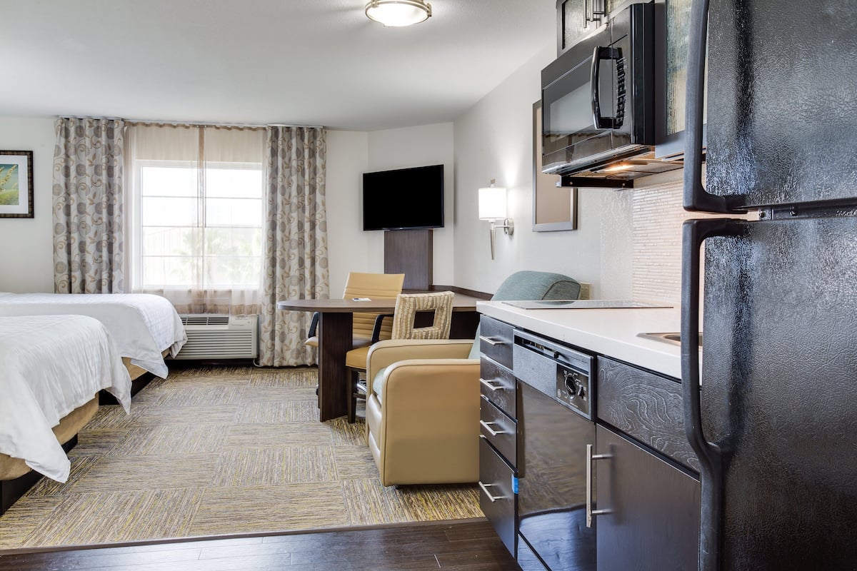 Une chambre d’hôtel avec deux lits, une table à manger avec des chaises, un coin cuisine avec un réfrigérateur et un téléviseur fixé au mur.