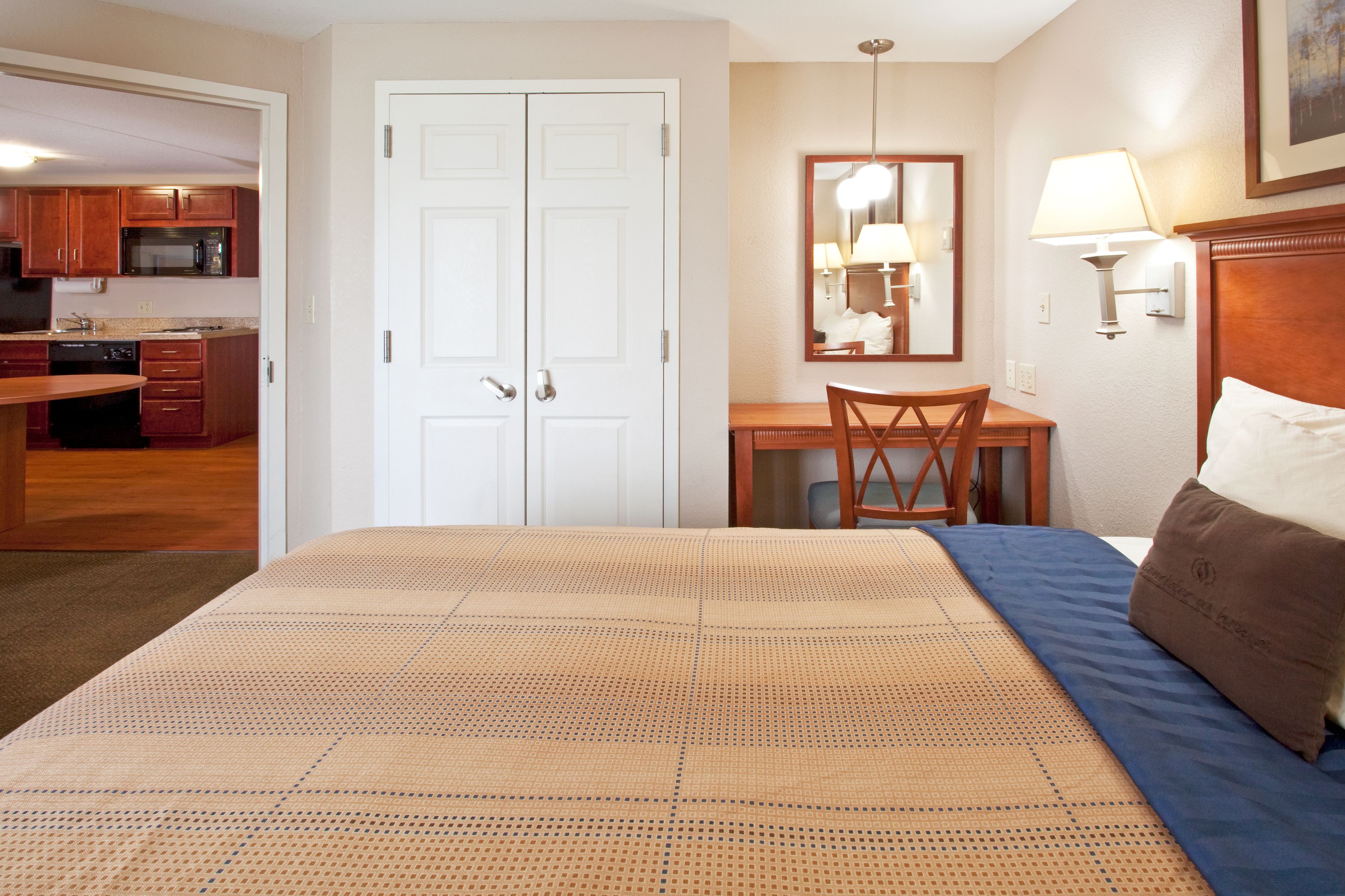 A hotel room with a bed, a desk, a chair, a dining table, and a kitchen area visible through an open door.