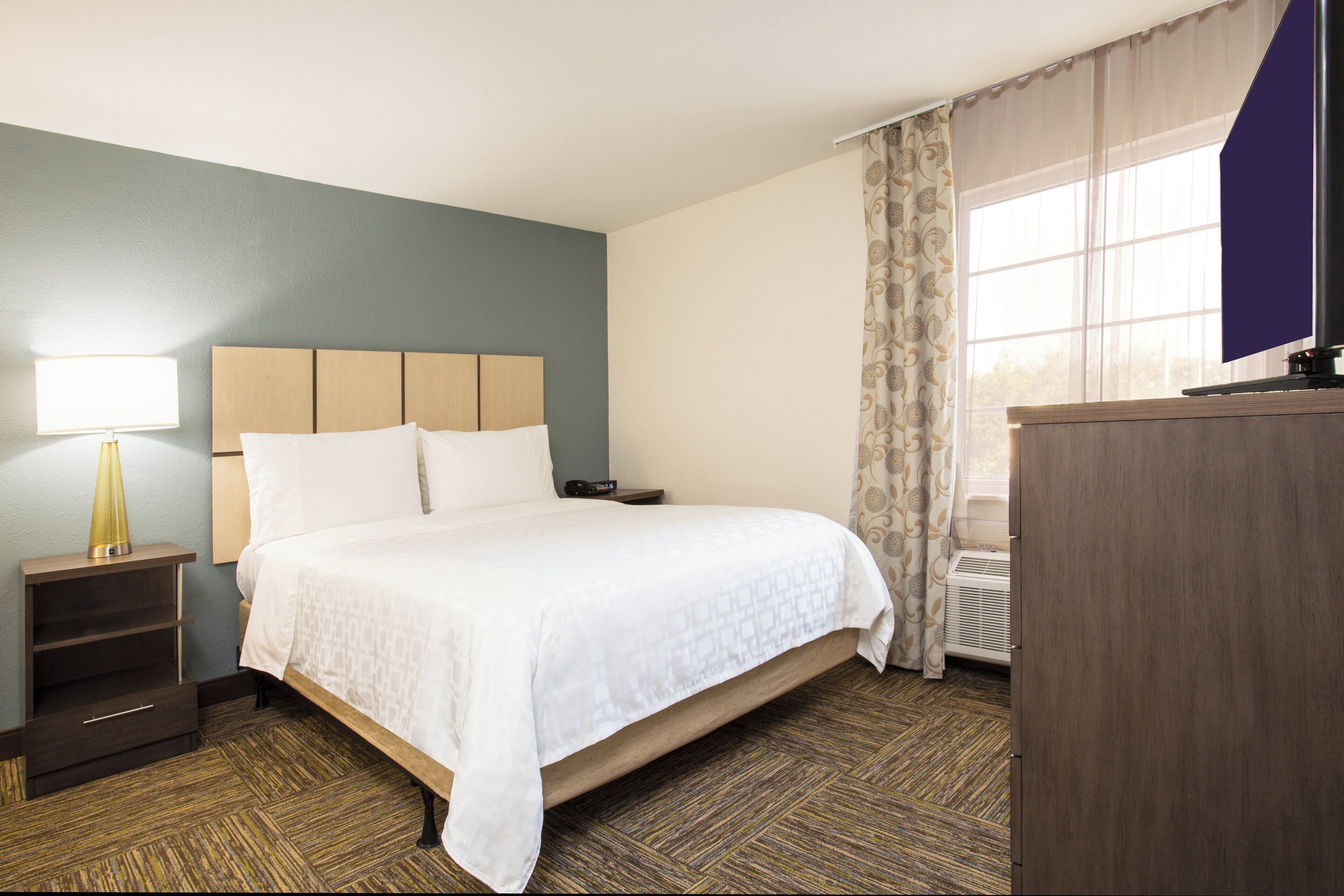 A hotel room with a bed, bedside table, lamp, and a window with curtains.