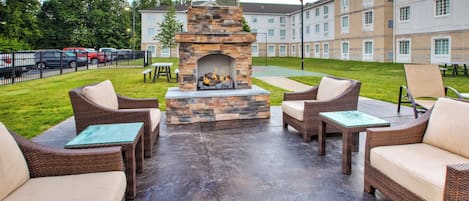 An outdoor patio area with wicker furniture, a stone fireplace, and a view of a multi-story building in the background.