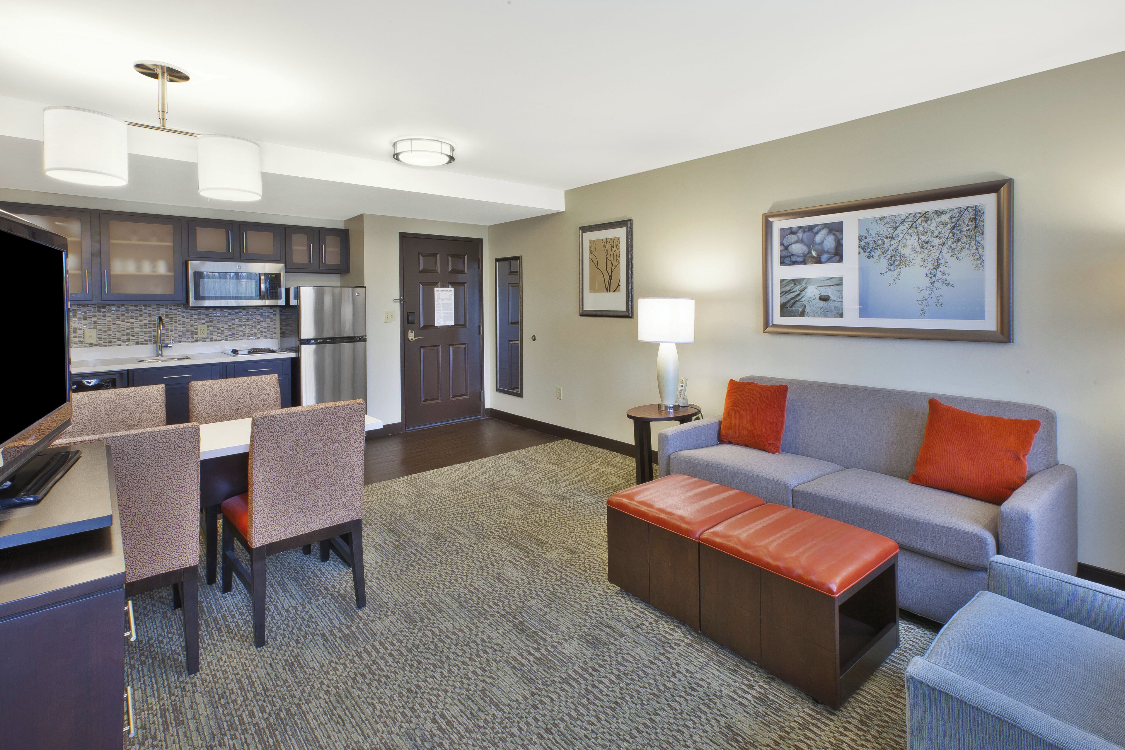 Una habitación de hotel moderna con kitchenette, comedor y sala de estar con sofá y reposapiés.
