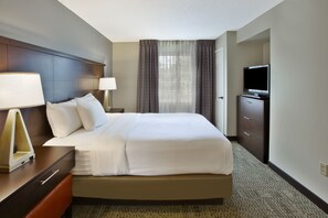 Quarto de hotel com uma cama grande, televisão, secretária com luminária e janela com cortinas.