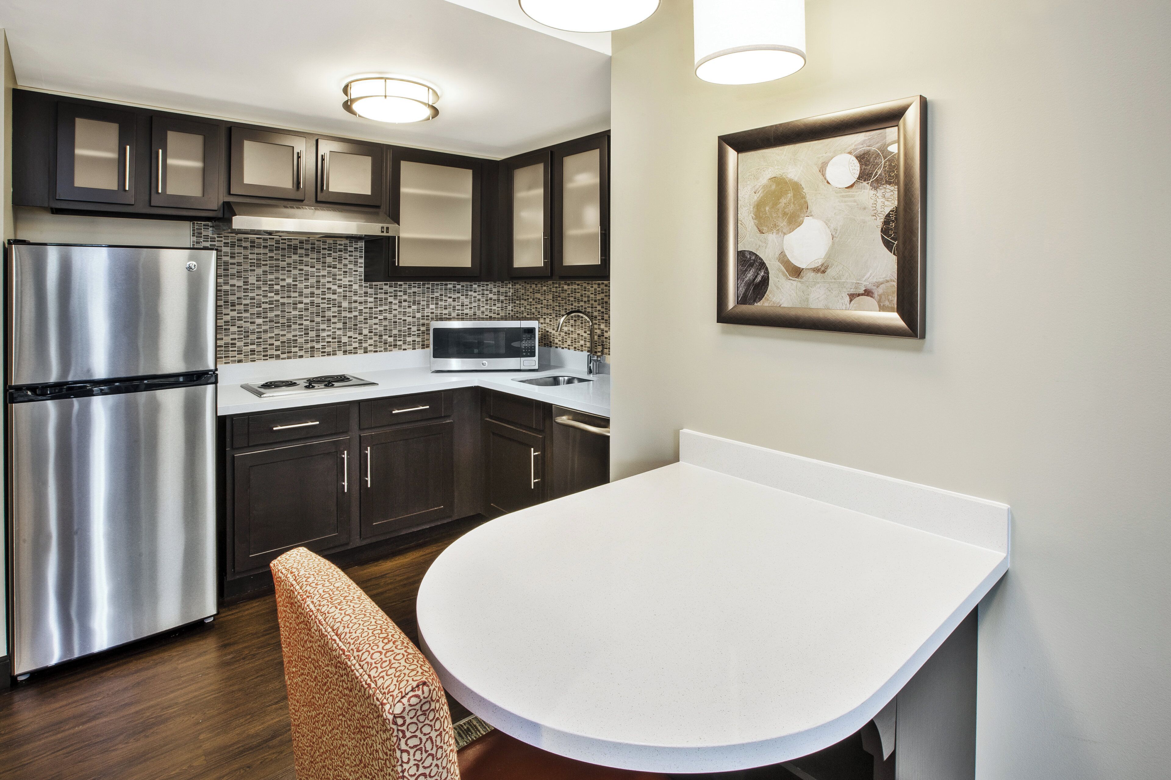A modern kitchen with a stainless steel refrigerator, a microwave, and a sink. There is a white countertop and dark cabinets.