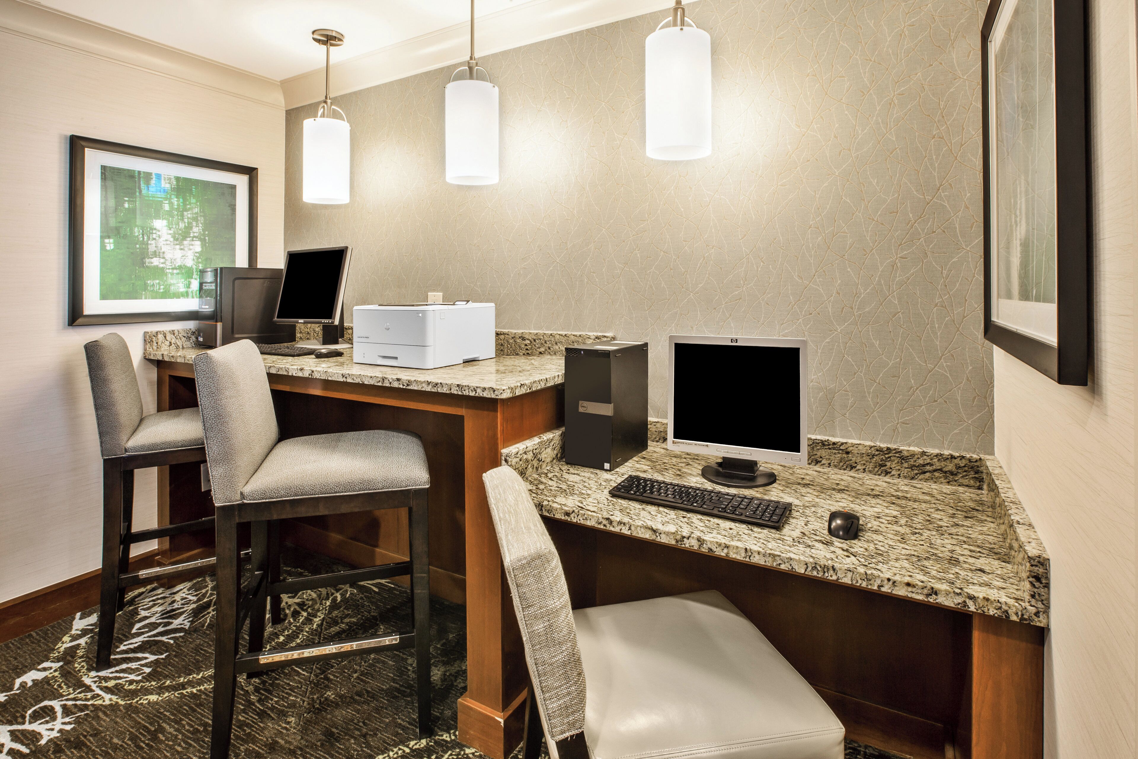 A hotel room with a desk, two chairs, a printer, two monitors, a keyboard, a mouse, and a framed picture on the wall.