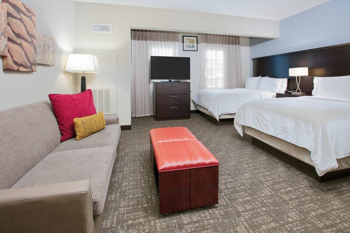 A hotel room with a sofa, two beds, a TV, a dresser, and a red ottoman.