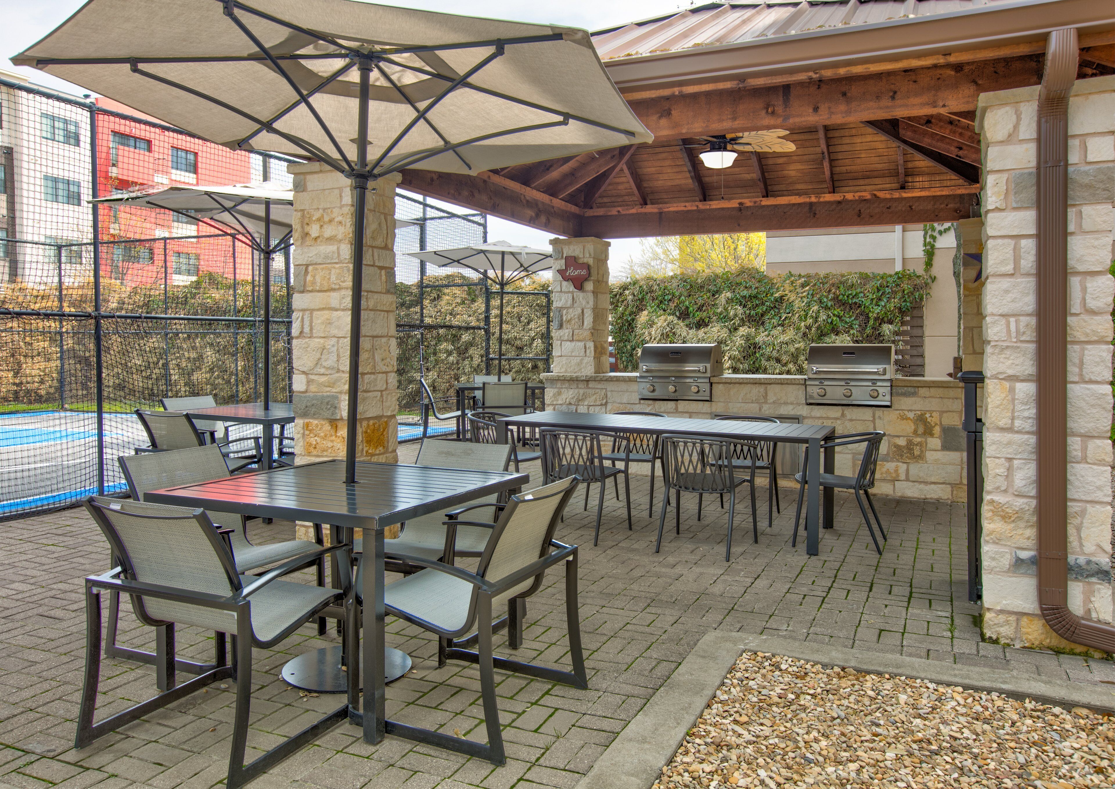 An outdoor patio with tables and chairs, a grill, and a pool in the background.