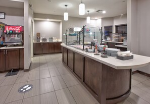 A modern kitchen area with a Coca-Cola dispenser, a buffet setup with glass display cases, and a clean tiled floor. - Staybridge Suites Plano - Richardson Area by IHG (Plano)