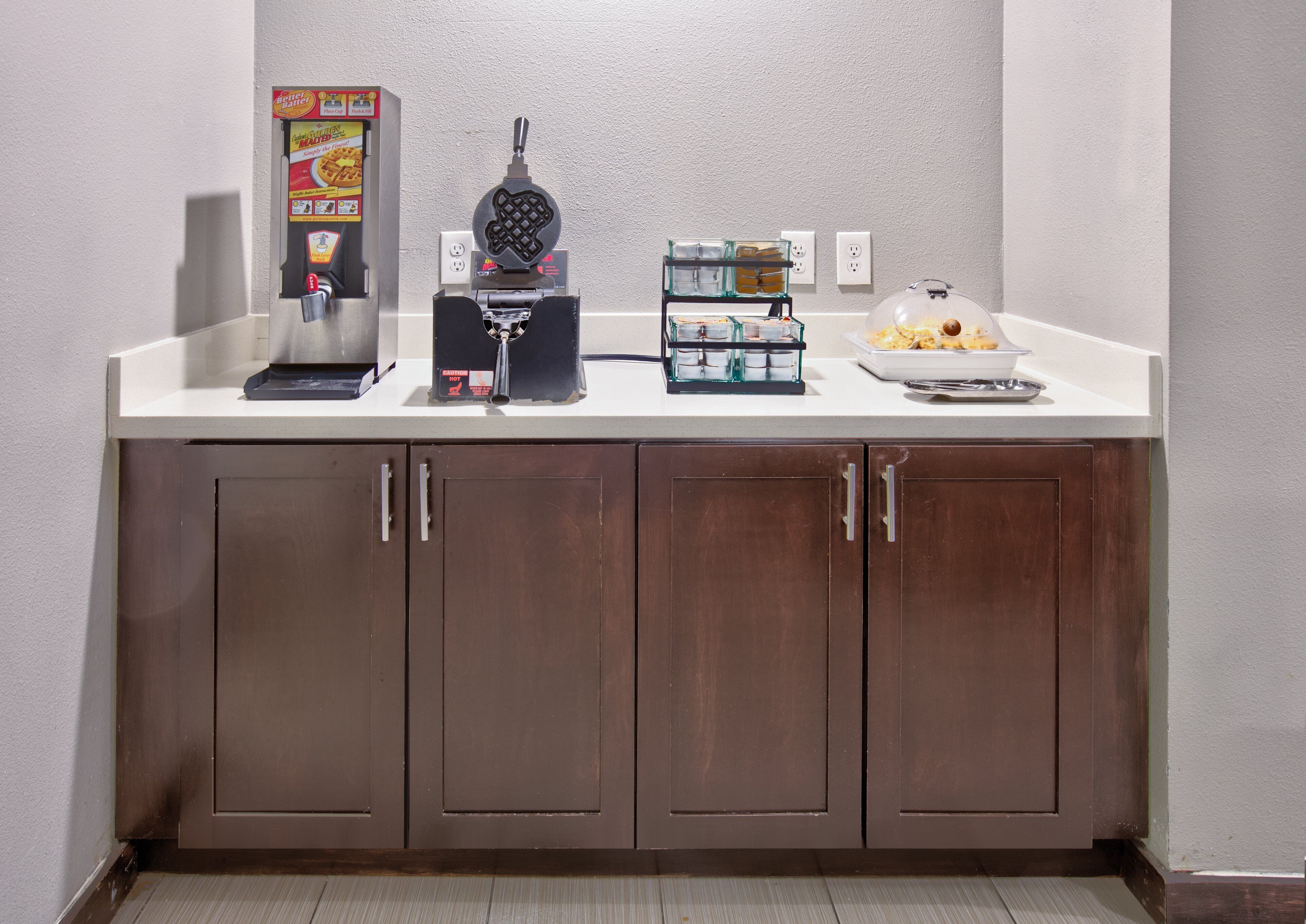 A kitchenette with a waffle maker, a coffee maker, and a glass container with utensils.
