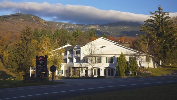 A white building with a signboard in front, surrounded by trees and mountains in the background. - The Inn at the Mountain & Condominiums (Stowe)