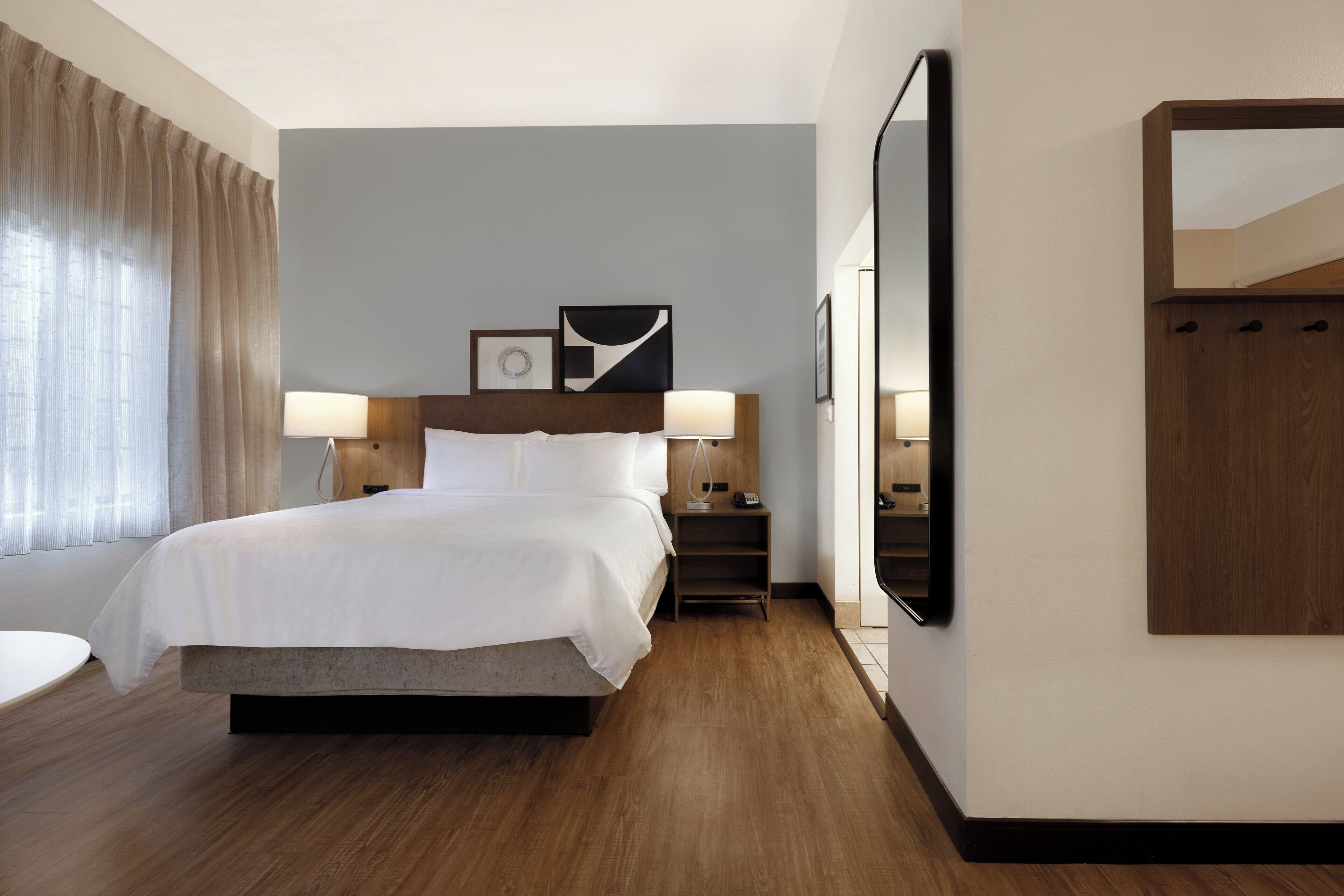 A hotel room with a bed, bedside tables, a mirror, and a window with curtains.