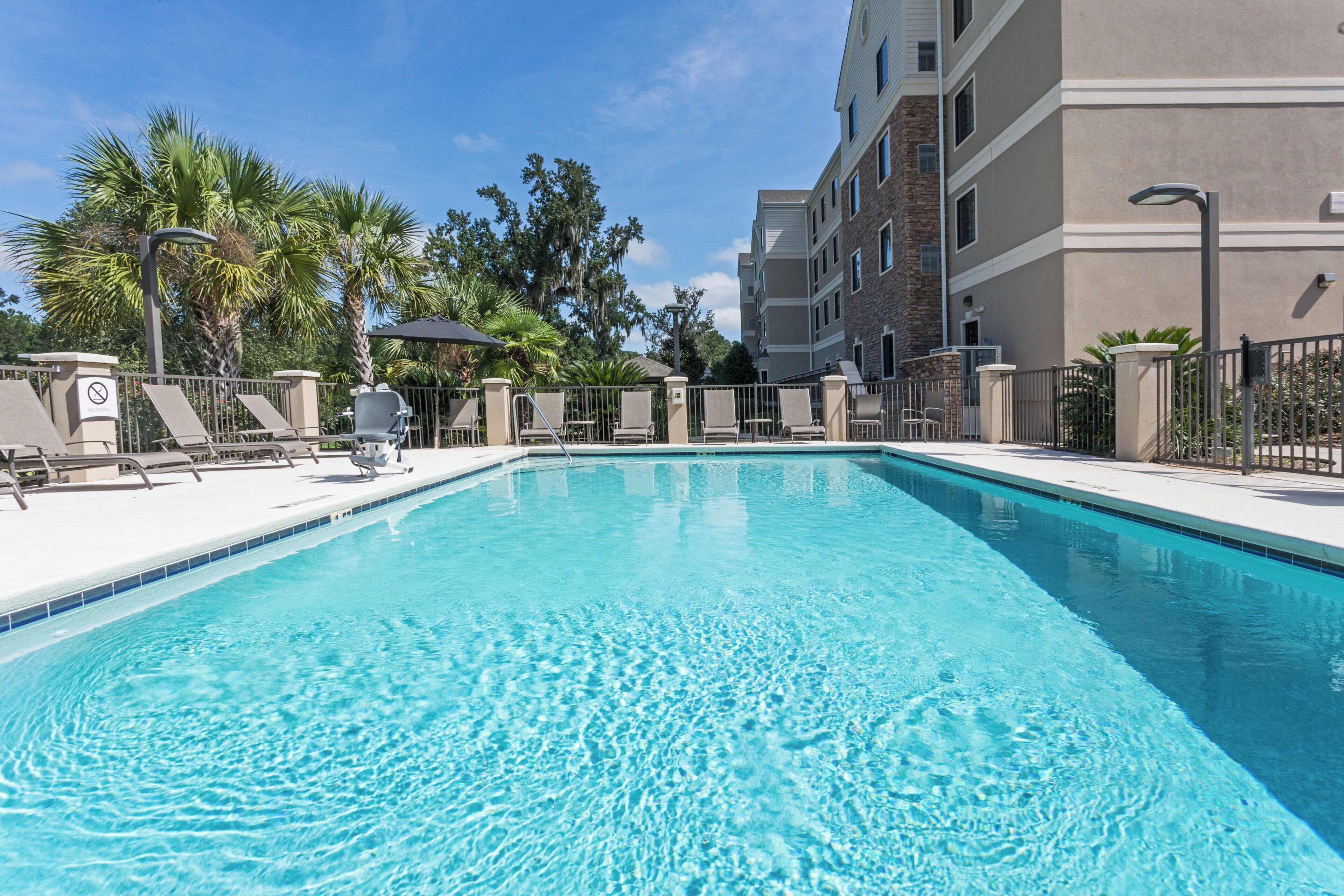 A clear blue pool with lounge chairs and umbrellas, surrounded by a fence and trees.