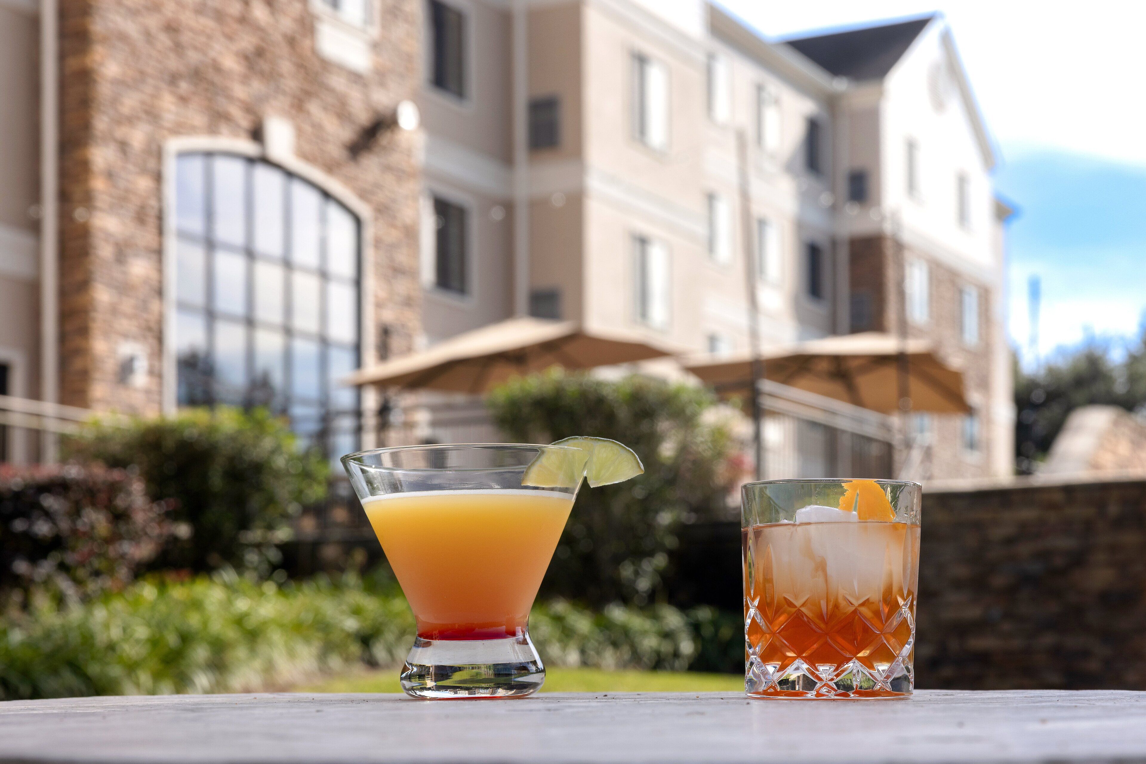 Deux boissons sur une table, avec un bâtiment en pierre et de la verdure en arrière-plan.