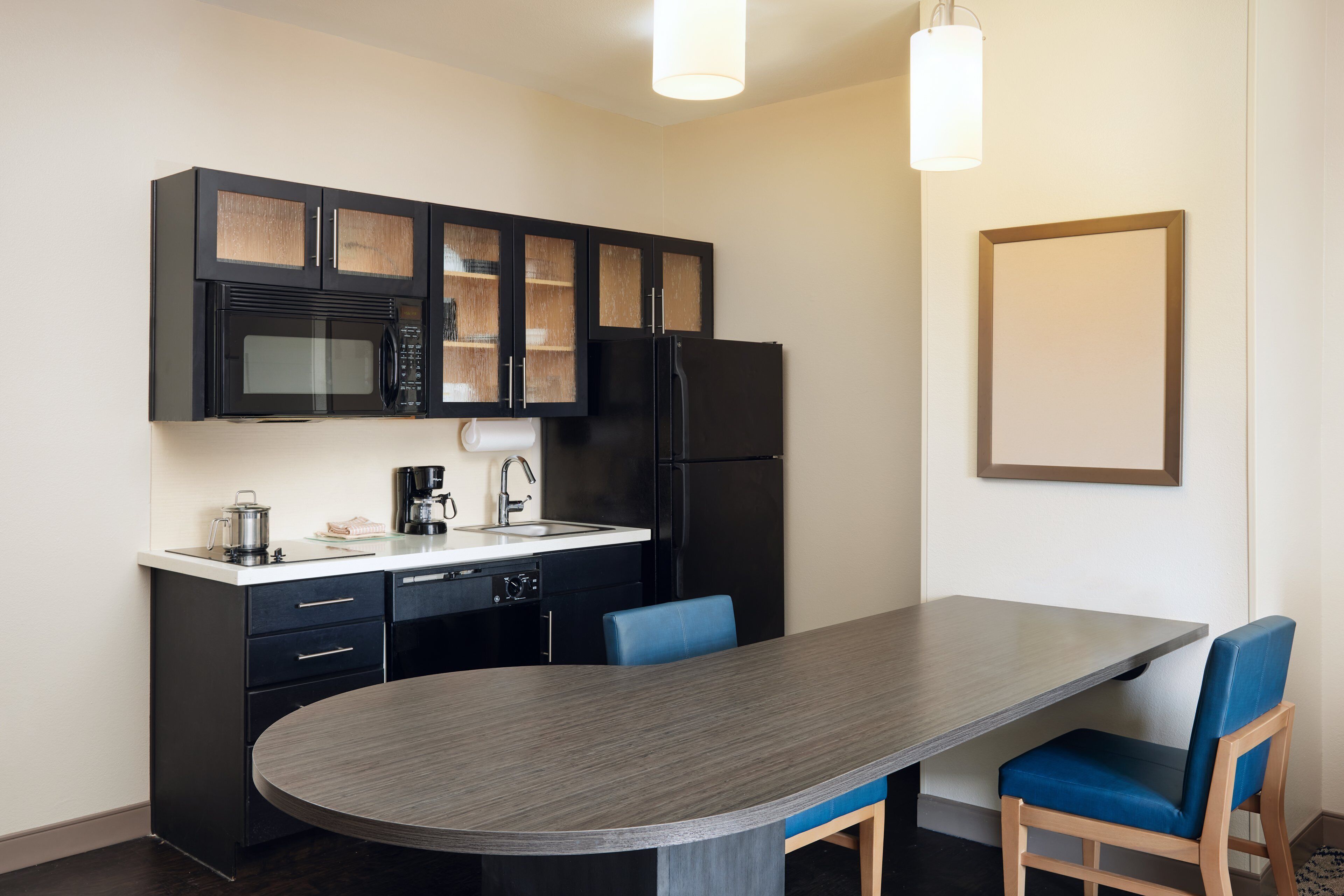 A compact kitchen with a microwave, refrigerator, and coffee maker. There is a dining table with blue chairs and a light fixture above.