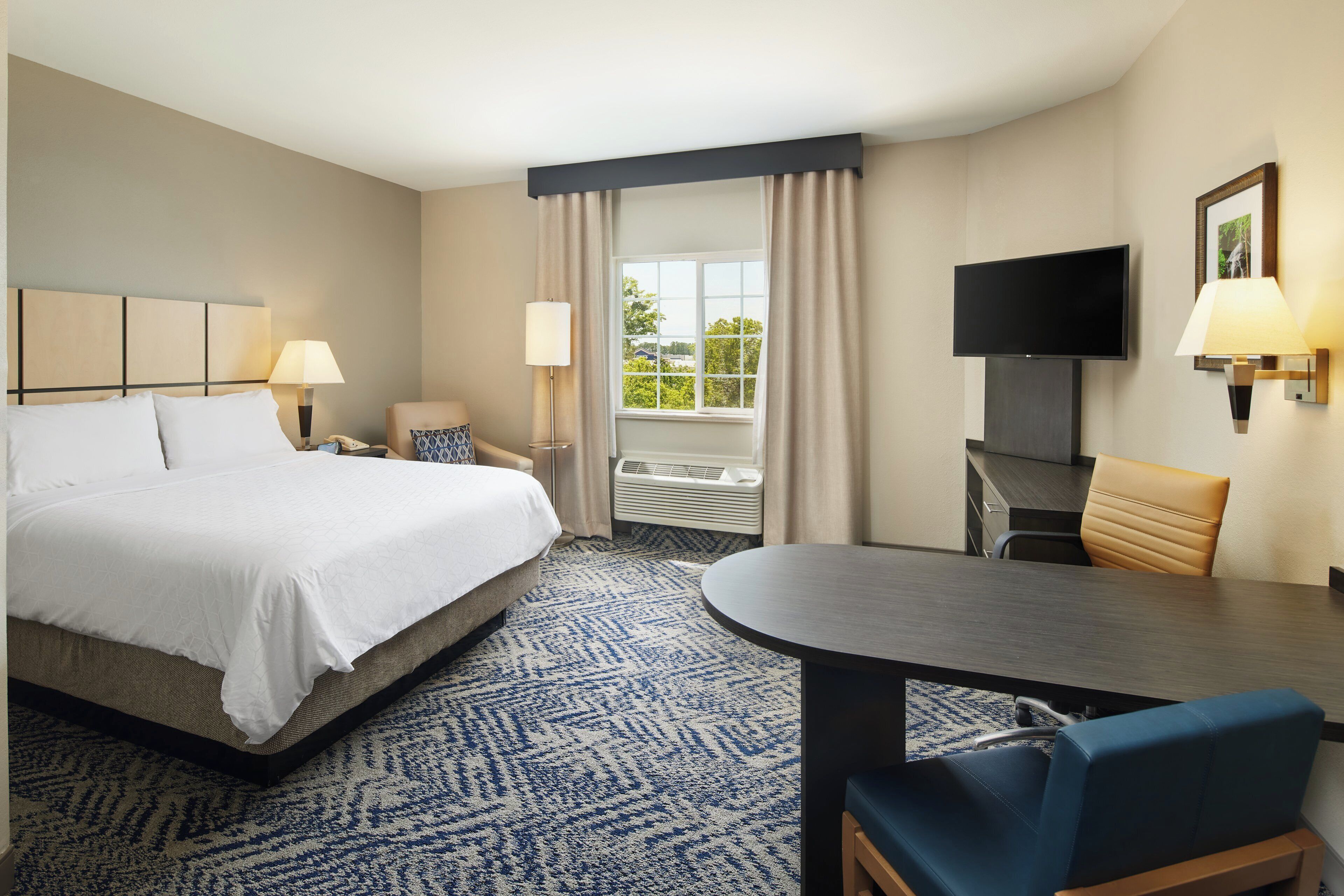 A hotel room with a bed, two bedside lamps, a television, a desk with a chair, and a large window with curtains.