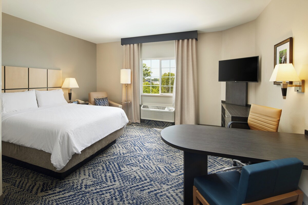 Quarto de hotel com cama, escrivaninha, cadeira, televisão e janela com cortinas.