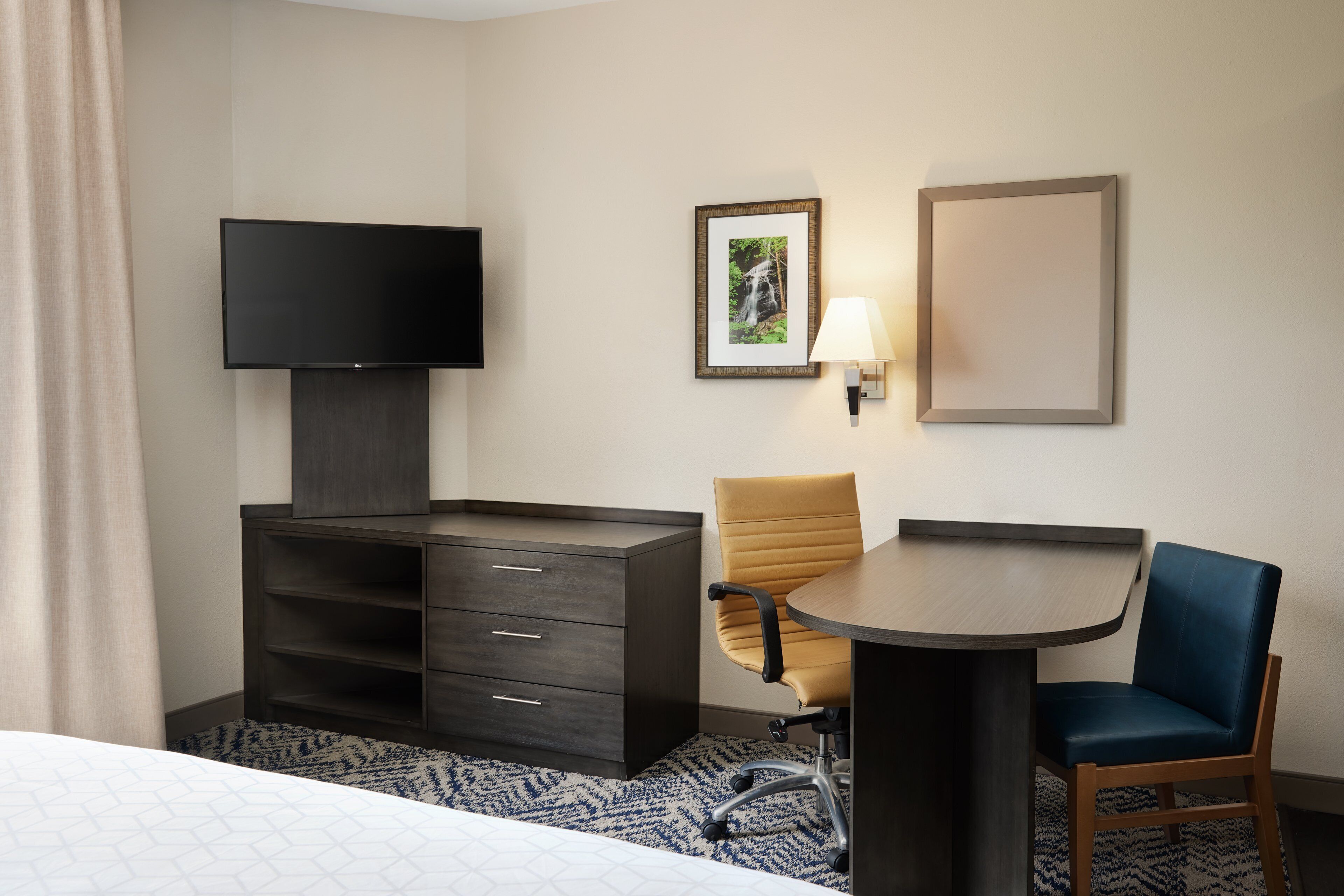 A hotel room with a bed, a TV, a desk with a chair, a dresser, and a wall-mounted lamp.