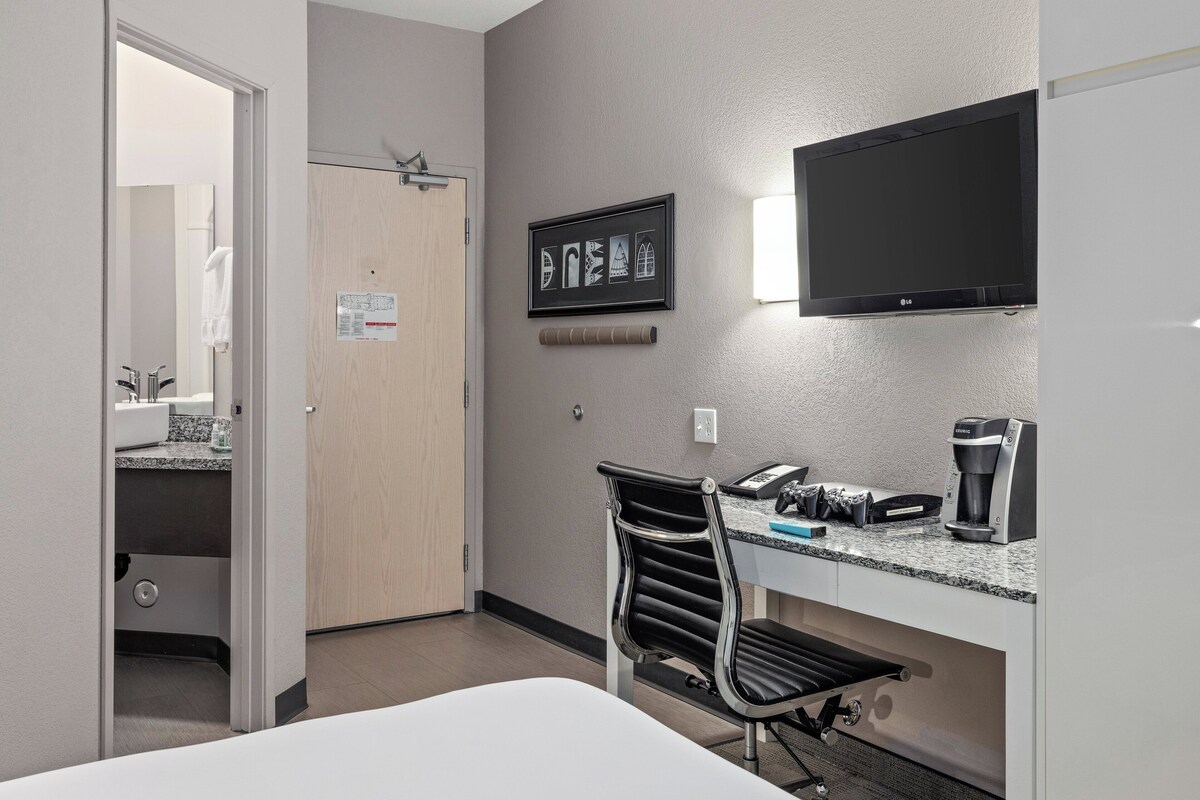 A hotel room with a bed, a desk with a coffee maker, a TV, and a chair.
