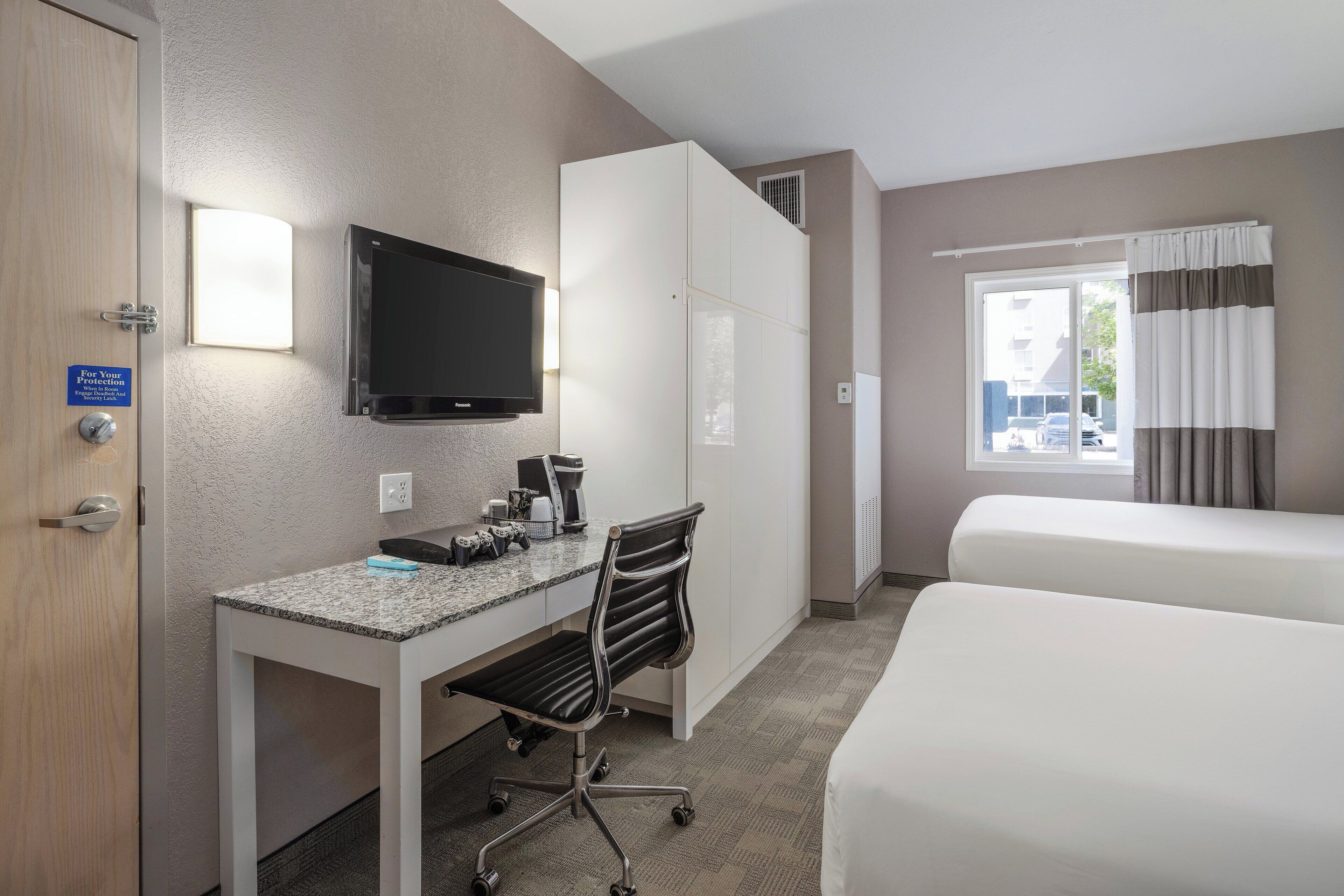 Habitación de hotel con escritorio, silla, televisor, cama y ventana con cortinas.