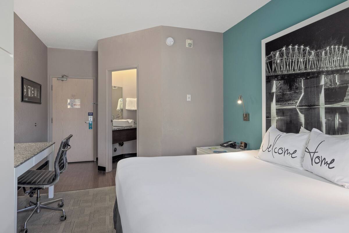 Une chambre d’hôtel avec un lit, un bureau, une chaise, un miroir et un tableau encadré au mur.