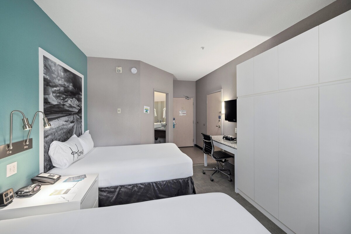 A hotel room with two beds, a desk, a chair, a mirror, and a large window.