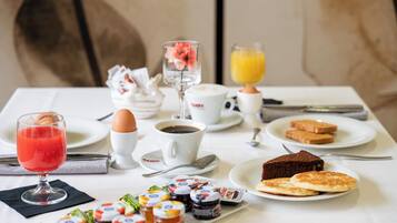 Une table de petit-déjeuner avec divers aliments, notamment des toasts, des crêpes, un verre de jus d’orange, une tasse de café et un verre de jus rouge.