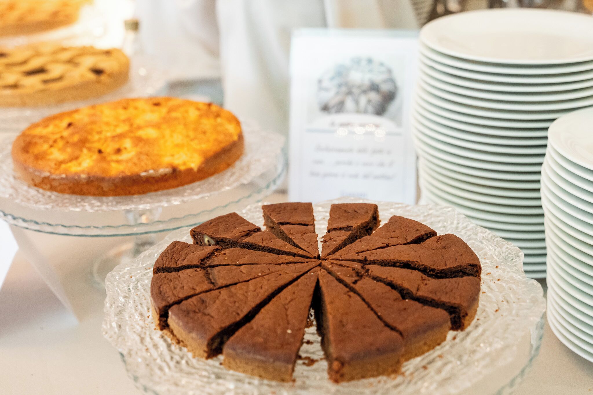 A dessert display with a chocolate cake and a pie on glass stands, surrounded by stacked plates.