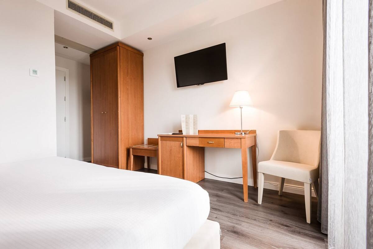 A hotel room with a bed, a wooden desk, a chair, a TV, and a lamp.