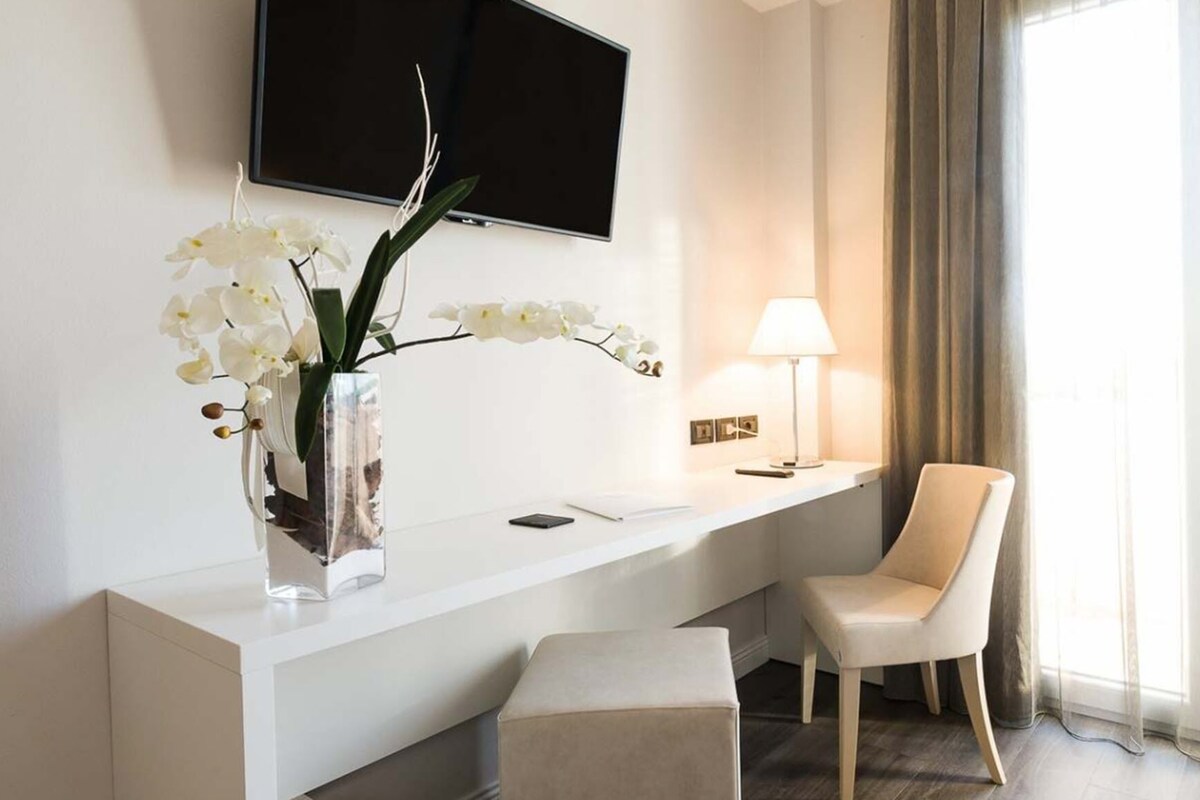 Una habitación de hotel con cama, escritorio con lámpara, silla y un jarrón con flores blancas.