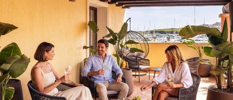 Three people seated at an outdoor table with food and drinks, surrounded by potted plants and wicker furniture, with a view of boats in the background.