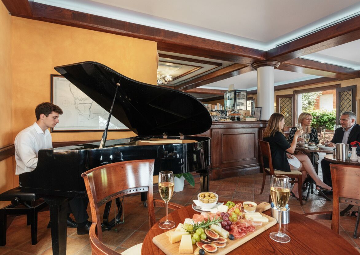 Een eetgedeelte met een piano, een tafel met een kaasplank, wijn glazen en mensen aan het dineren.