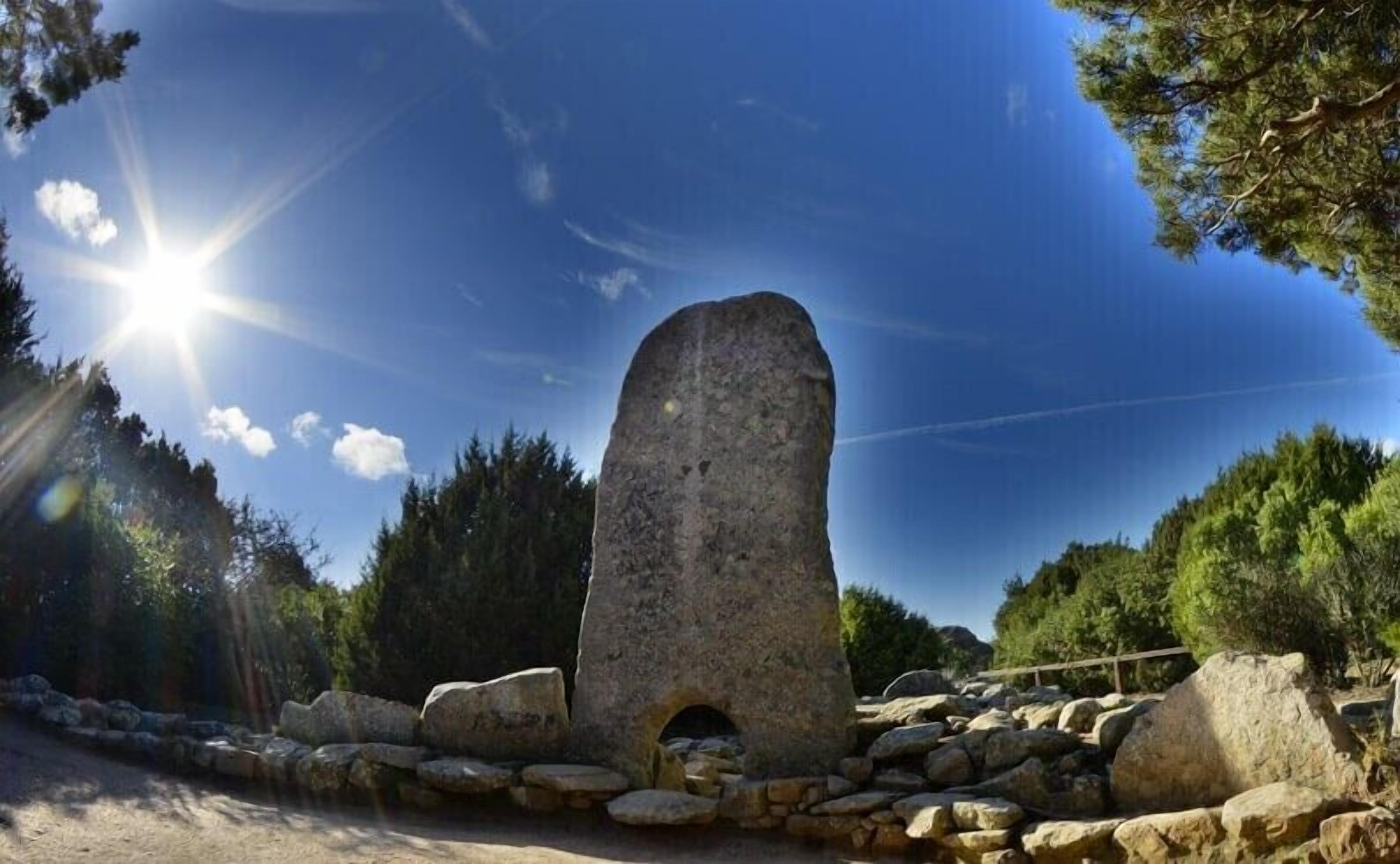 A stone structure with a central opening, surrounded by rocks and trees, under a clear sky.