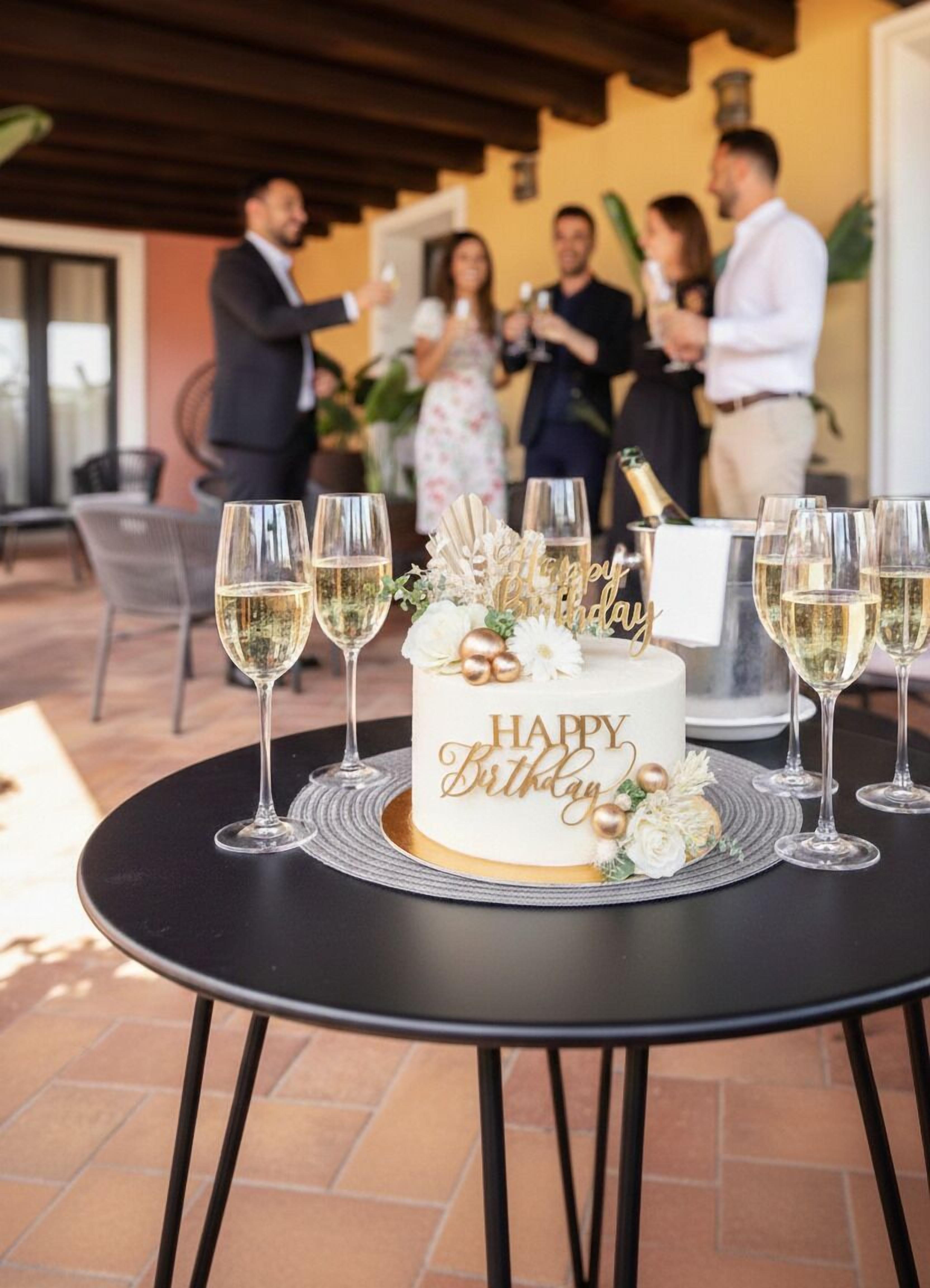 A round table with a birthday cake, surrounded by glasses of white wine, set outdoors.