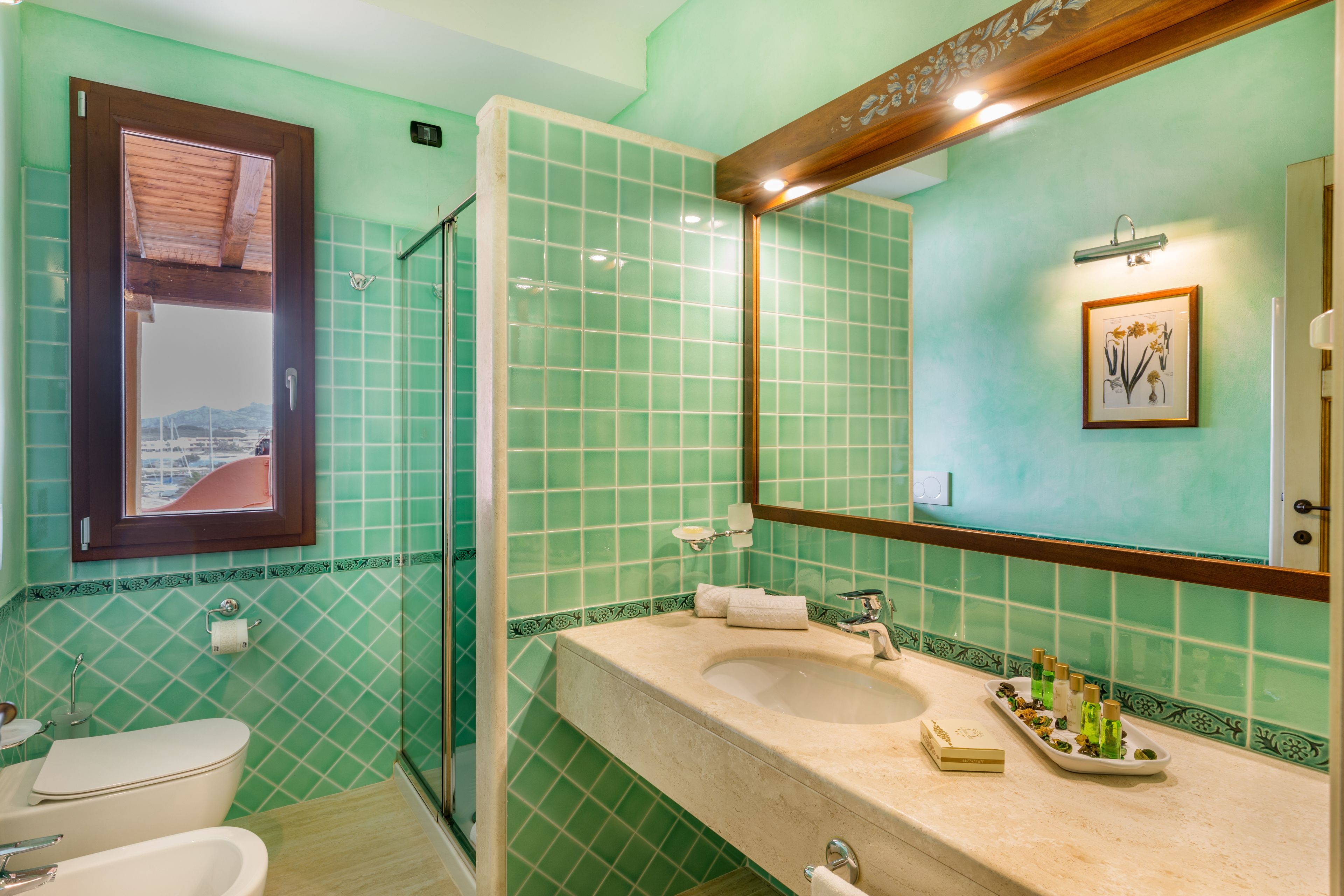 Un baño con pared de azulejos verdes, un lavamanos con artículos de aseo y un espejo.