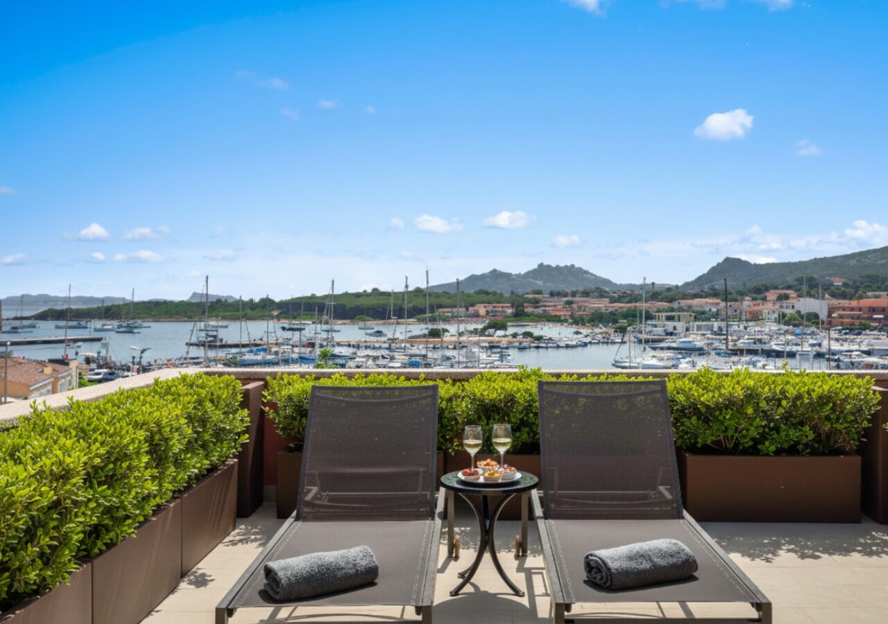 A terrace with two lounge chairs, a small table with a bottle and glasses, and a view of a marina with boats and buildings in the background.