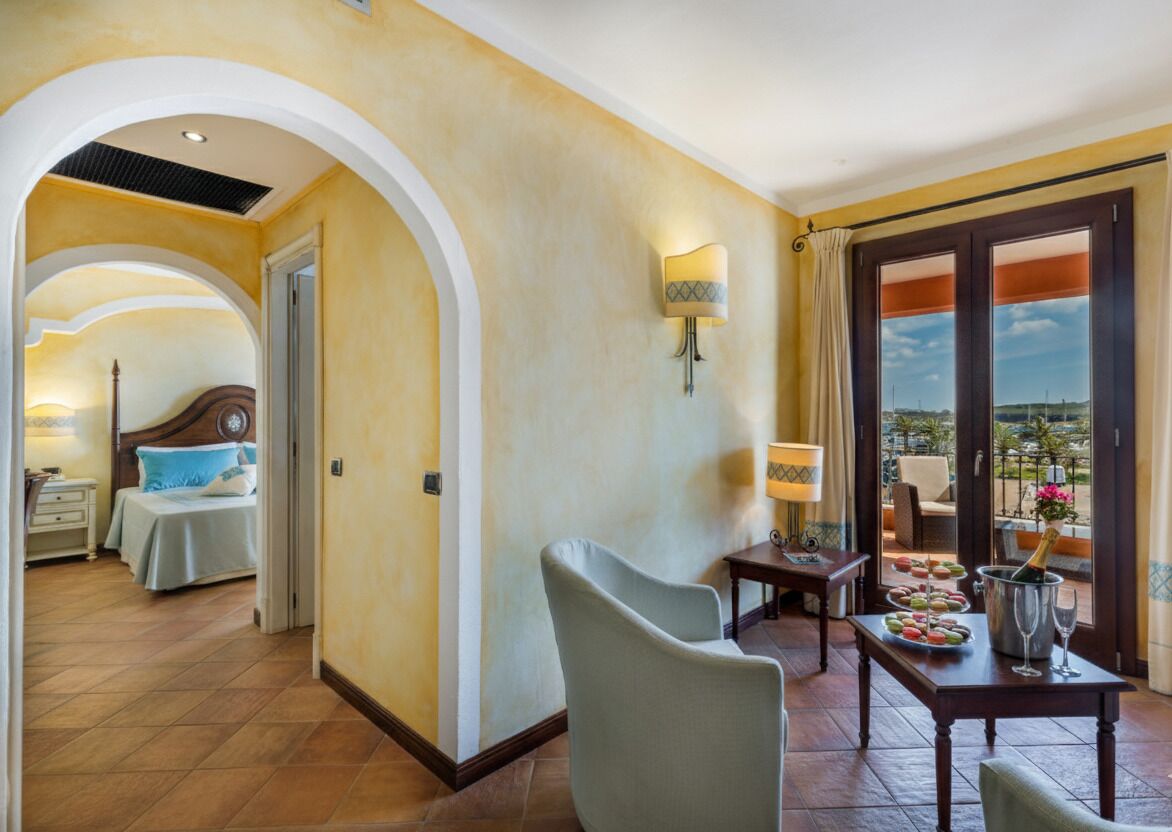 Quarto de hotel com cama, área de estar com mesa e cadeiras, varanda com vista e porta que dá para outro quarto.