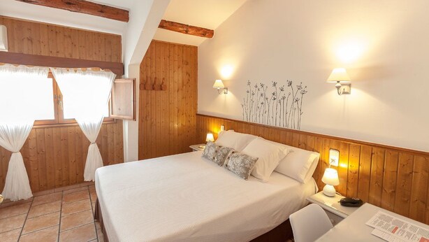 Quarto com uma cama grande, paredes de madeira, uma janela com cortinas e duas luminárias fixadas na parede.