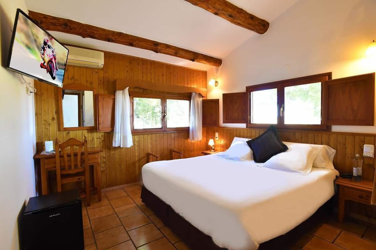 A bedroom with a large bed, wooden walls, a TV mounted on the wall, a desk with a chair, and a small table with a lamp.