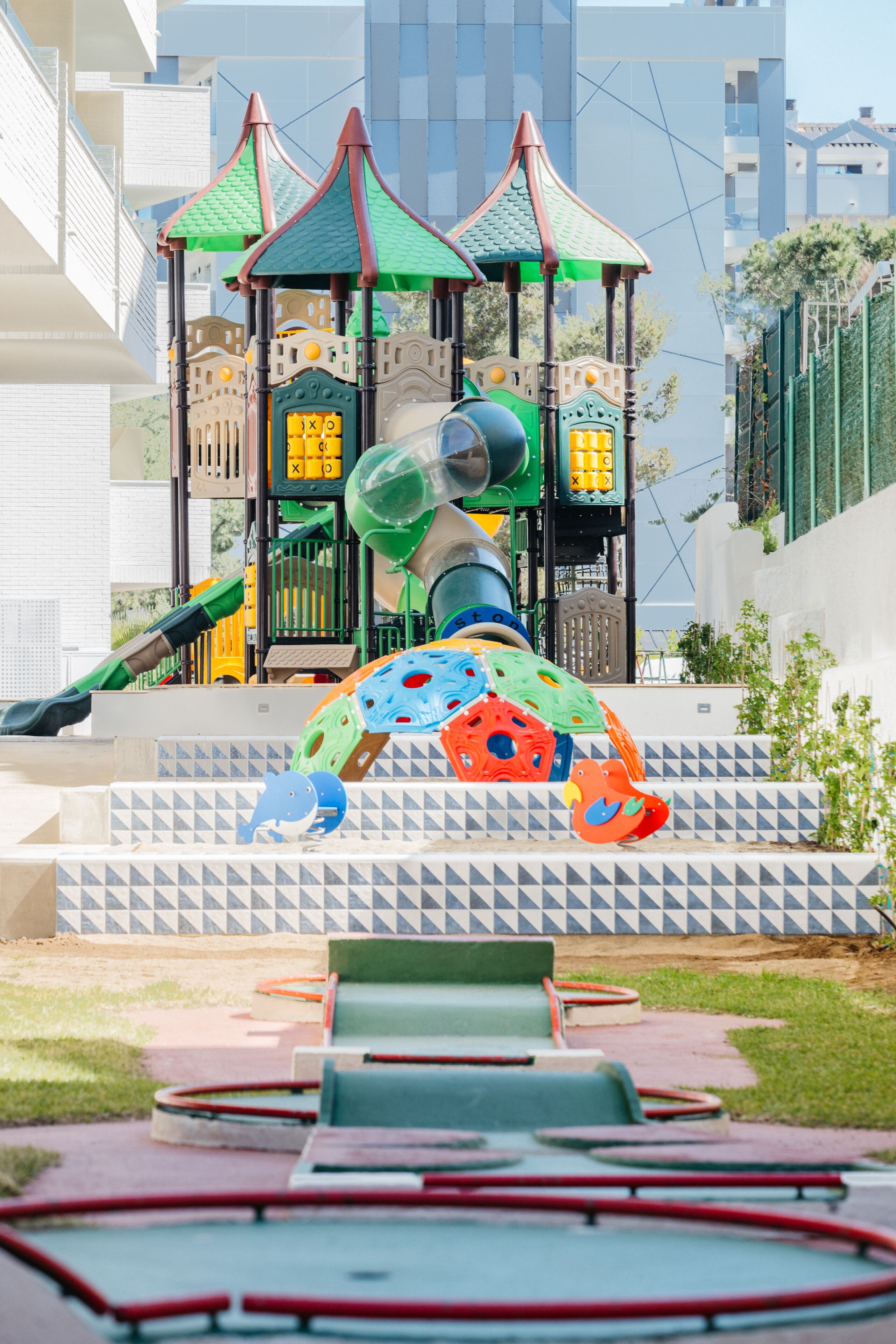 A playground with a climbing structure and slides, surrounded by a paved area with benches.