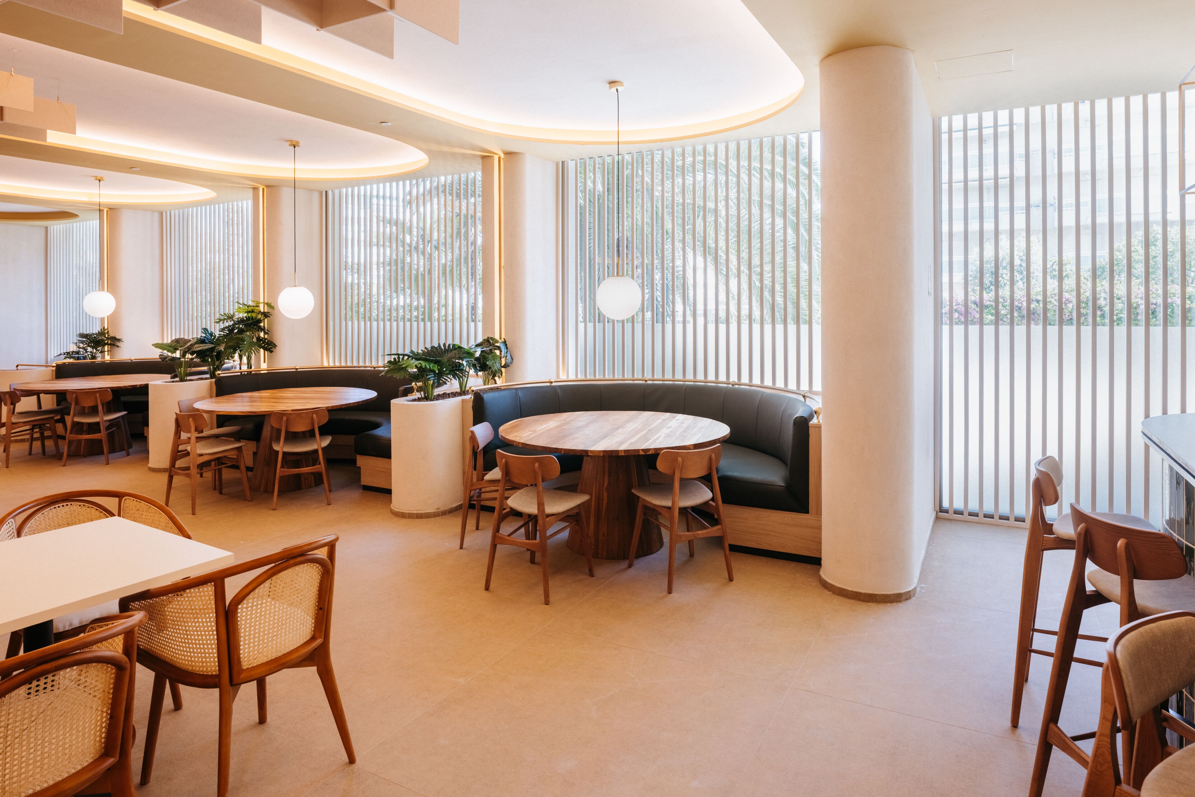 Un intérieur moderne avec des tables rondes, des chaises en bois, un banc incurvé et de grandes fenêtres équipées de stores verticaux.
