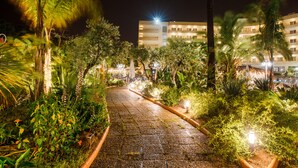 Un sendero nocturno bordeado de vegetación e iluminado por farolas, que conduce a un edificio de varios pisos y ventanas.