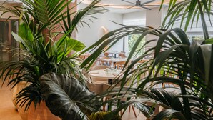 Un interior moderno con plantas en macetas grandes, suelo de madera y un área de comedor con mesas y sillas.