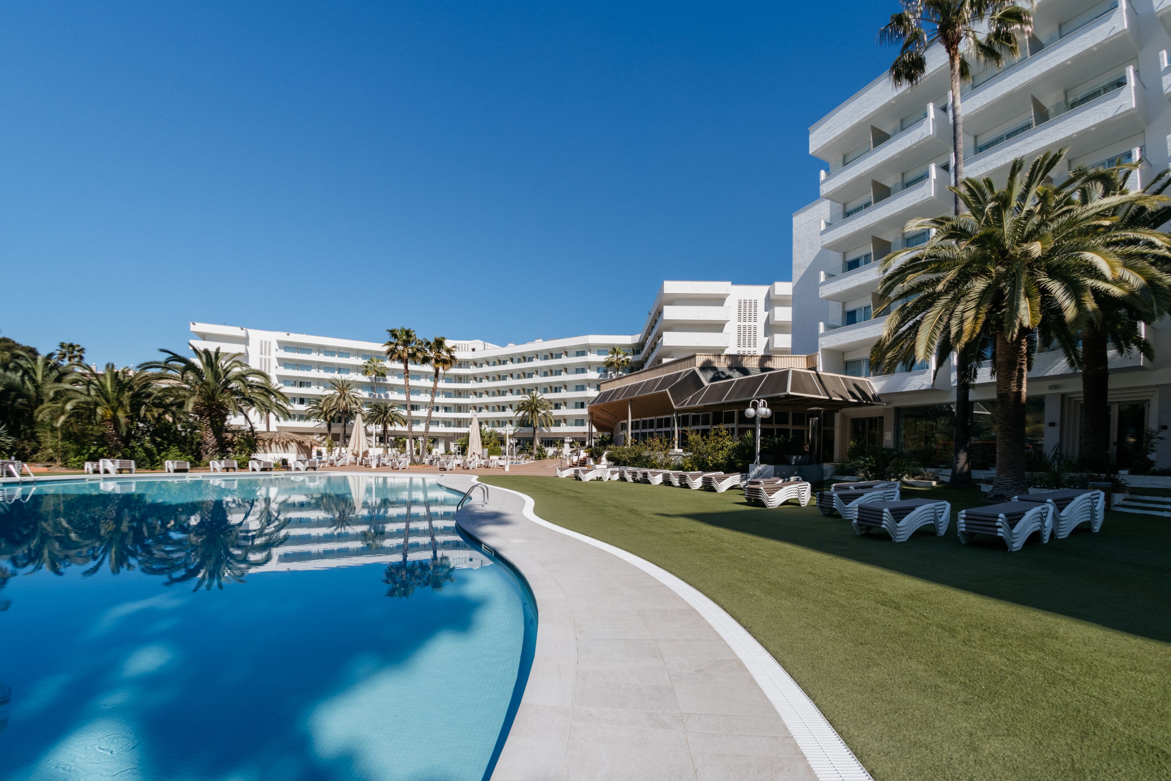 Una piscina con lettini, palme e un edificio di un hotel sullo sfondo.