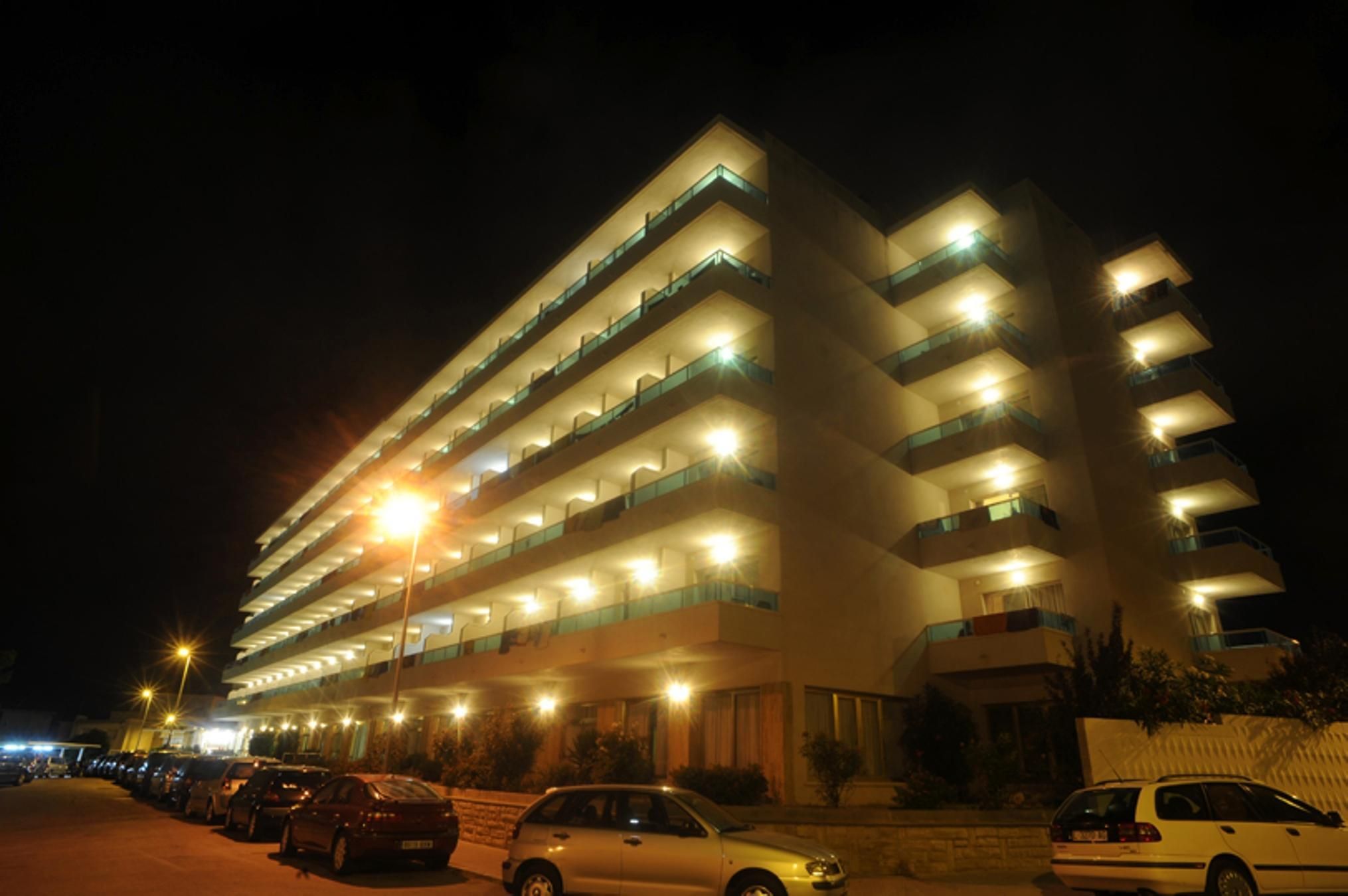 A multi-story hotel with illuminated balconies and a parking area in front.