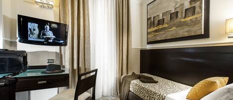 A hotel room with a bed, a desk with a laptop, a chair, a television on the wall, and a painting above the bed.