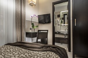 A modern hotel room with a bed, a desk with a chair, a potted plant, a flat-screen TV, and a large mirror.